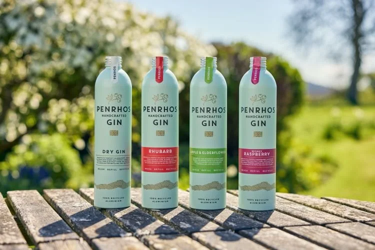 Four Penrhos Spirits aluminium gin bottles in Dry Gin, Rhubarb, Apple & Elderflower and Wonky Raspberry varieties displayed on a wooden outdoor table with a green orchard landscape in the background.