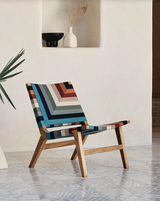Colorful woven lounge chair from MasayaCo with a geometric pattern on a natural wood frame in a bright, minimalist interior