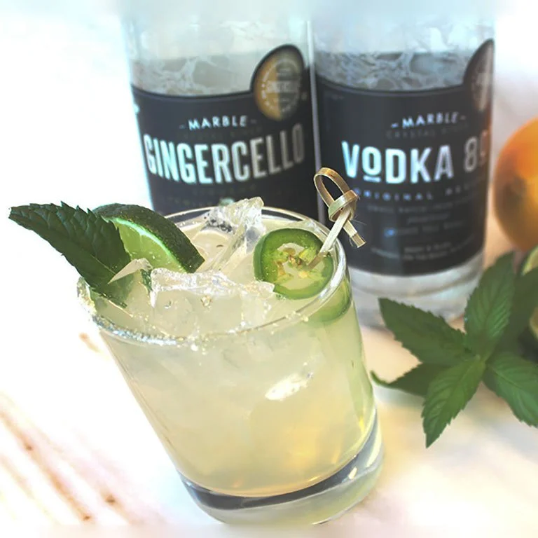 A cocktail with lime, mint and jalapeño garnish served over ice in front of Marble Distilling Gingercello and vodka bottles.