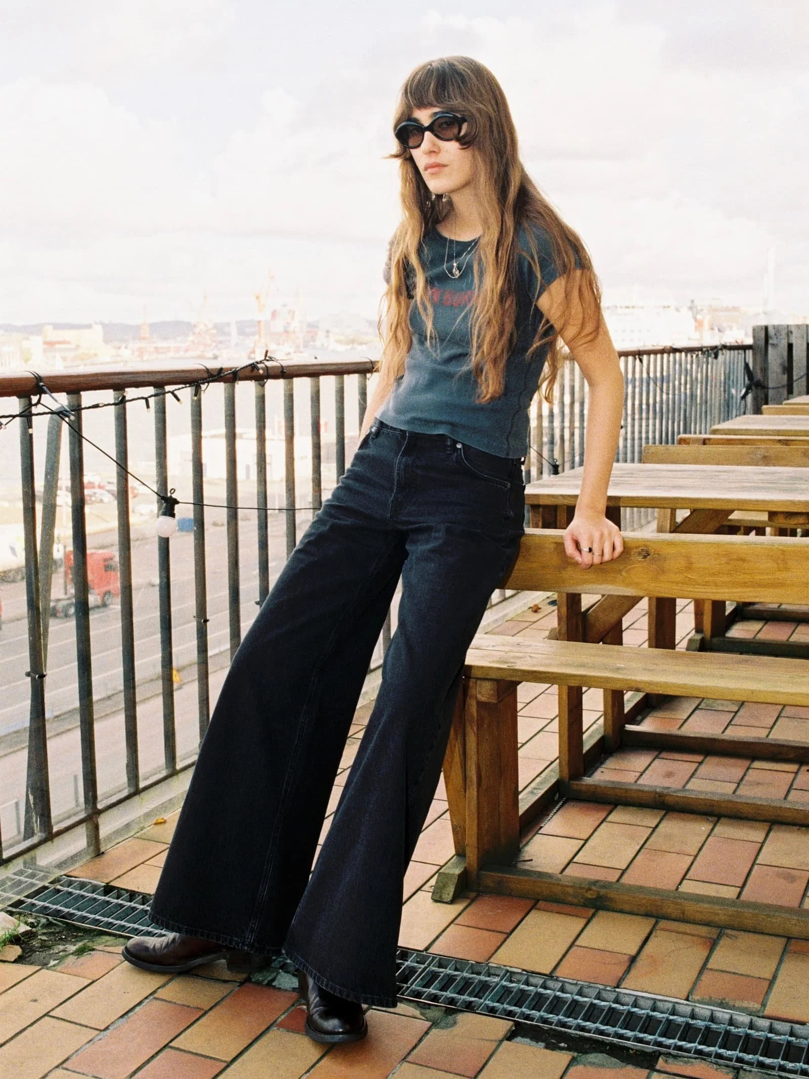 Person wearing dark wide-leg Nudie Jeans leaning against a wooden bench on a rooftop terrace, styled with a fitted tee, sunglasses and boots, with a cityscape in the background