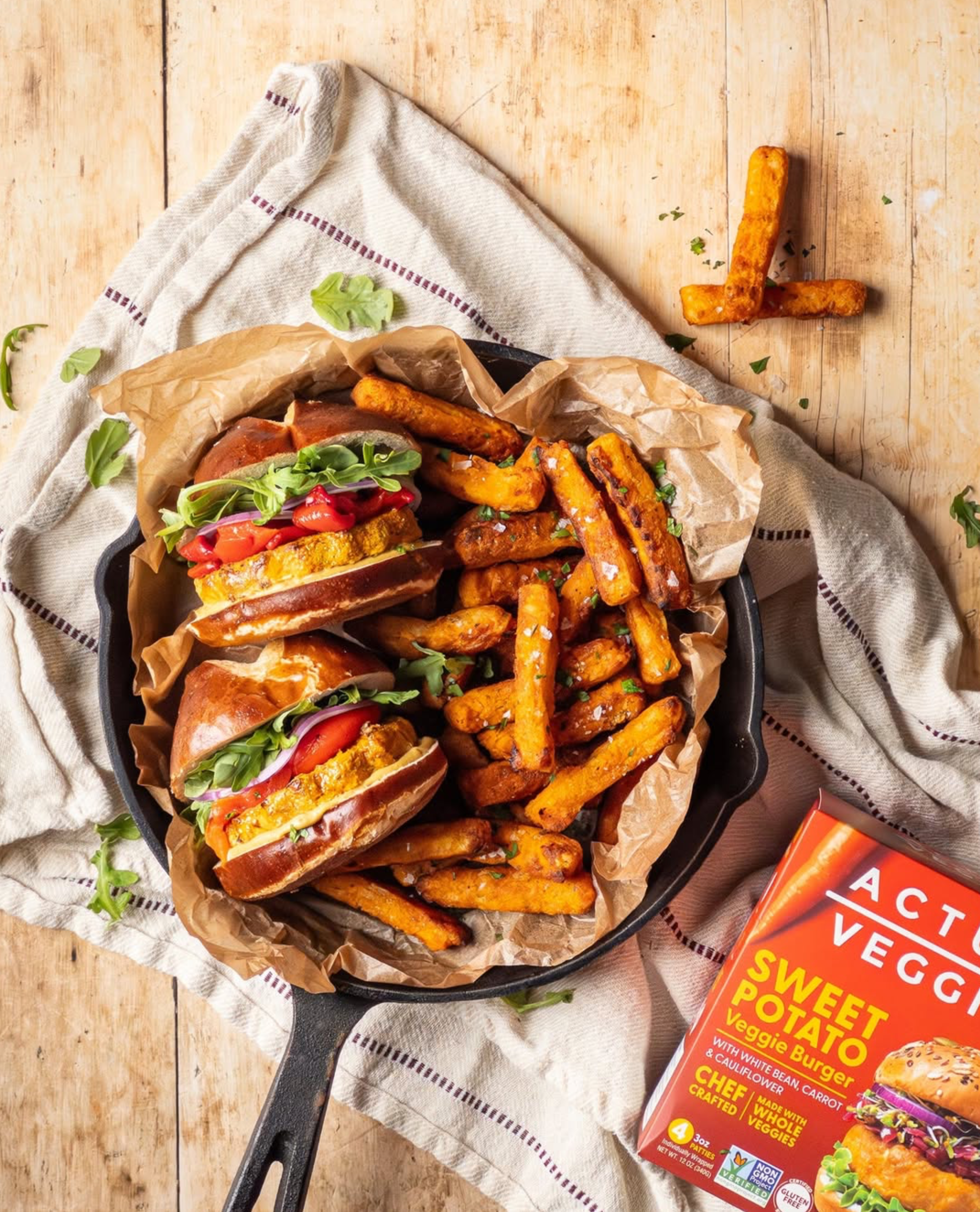 Two Actual Veggies Sweet Potato Veggie Burgers with white bean, carrot and cauliflower served on pretzel buns with arugula, red onion and tomato, alongside sweet potato fries in a cast iron skillet.