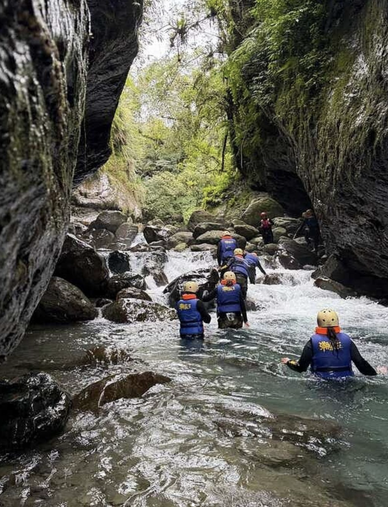 Group of people canyoning through a rocky river gorge, wearing helmets and life vests as they wade through flowing water surrounded by steep, mossy cliffs