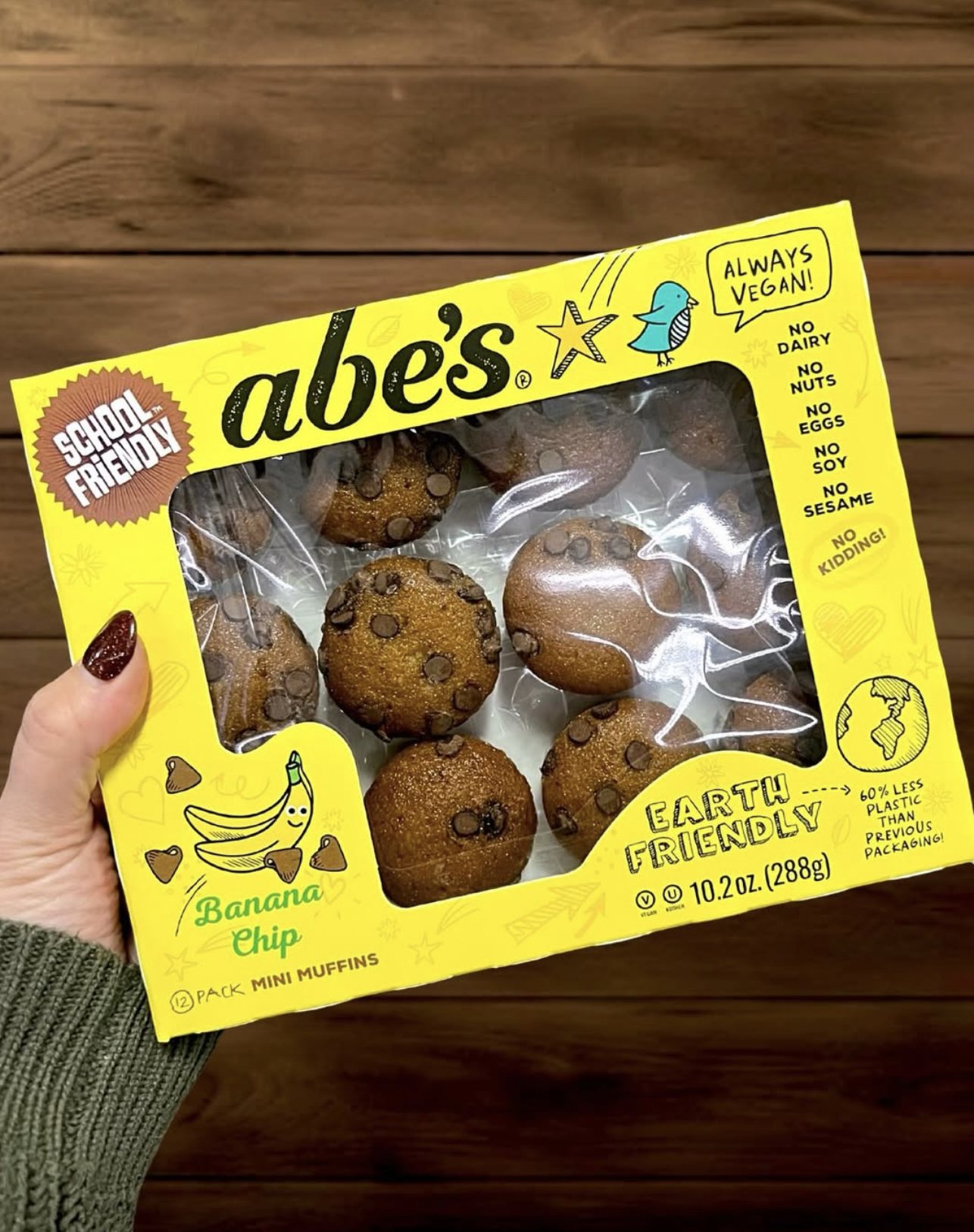 Hand holding a box of Abe’s Banana Chip Mini Muffins in bright yellow School-Friendly packaging featuring Earth Friendly and Always Vegan callouts.