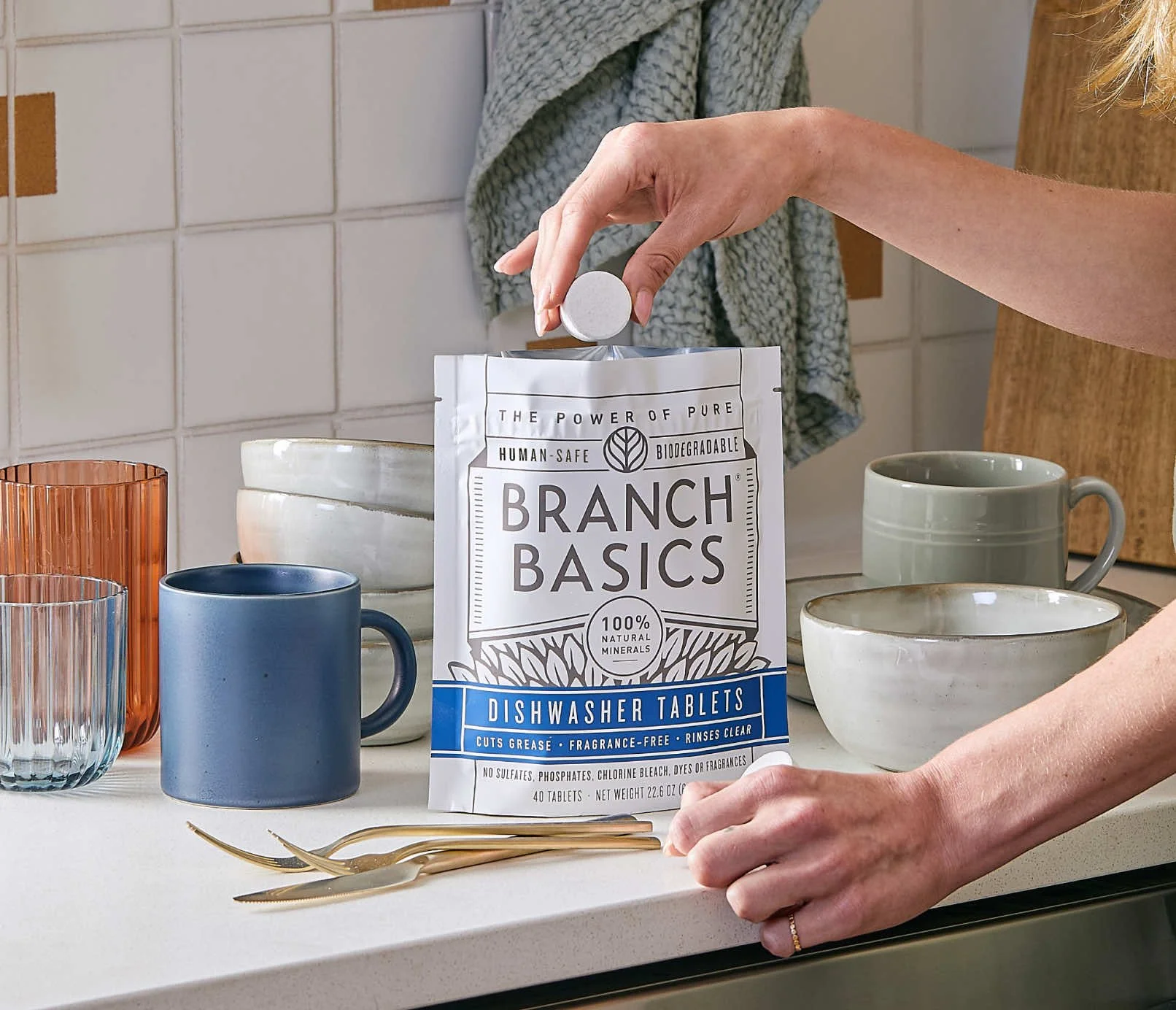 Hand placing a Branch Basics dishwasher tablet into a refill pouch beside clean dishes, highlighting a human-safe, low-waste dishwashing routine