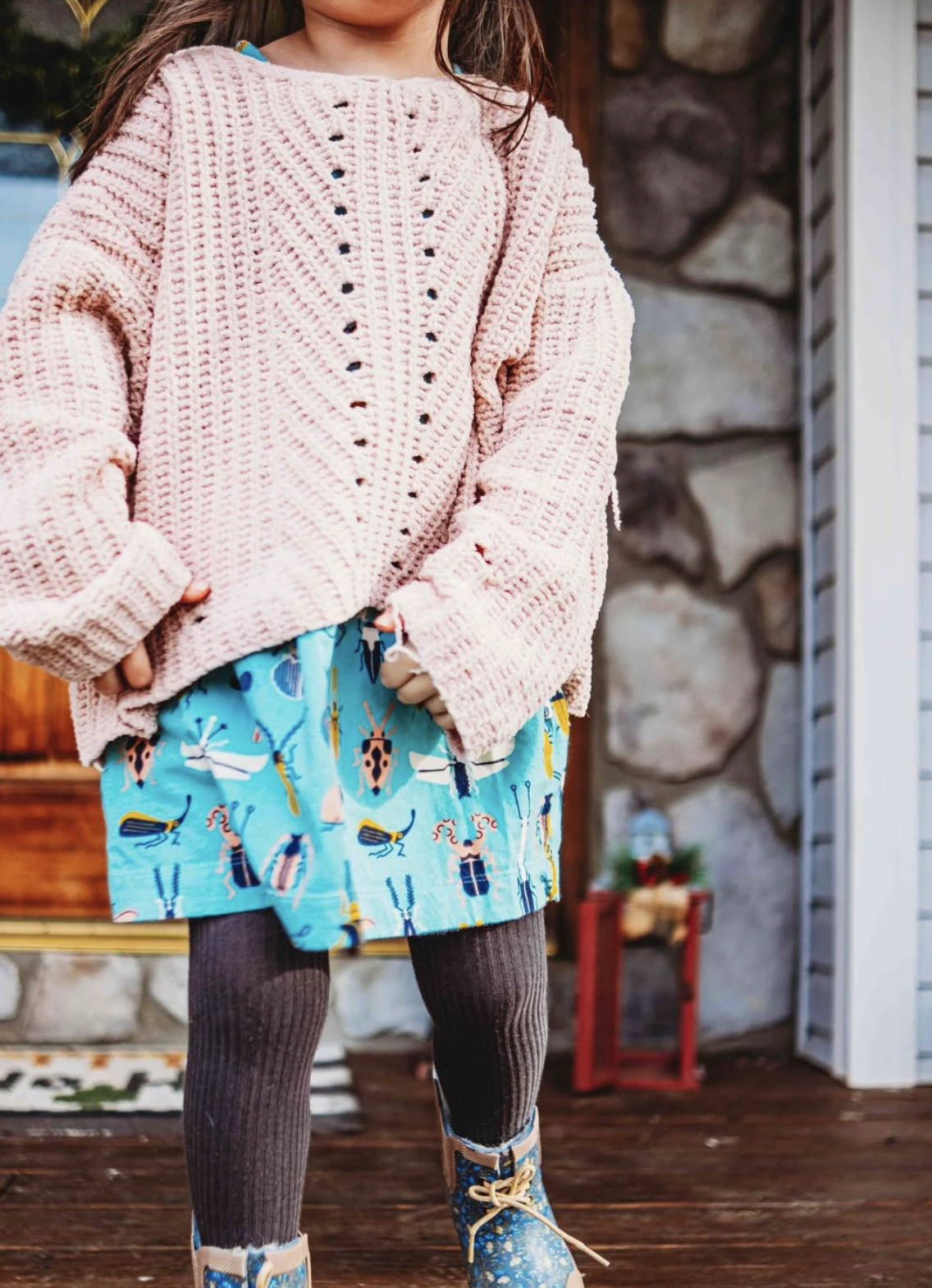 Child standing on a porch wearing a chunky knit sweater, printed skirt with insects, ribbed tights and lace-up boots, highlighting durable, playful kids clothing.