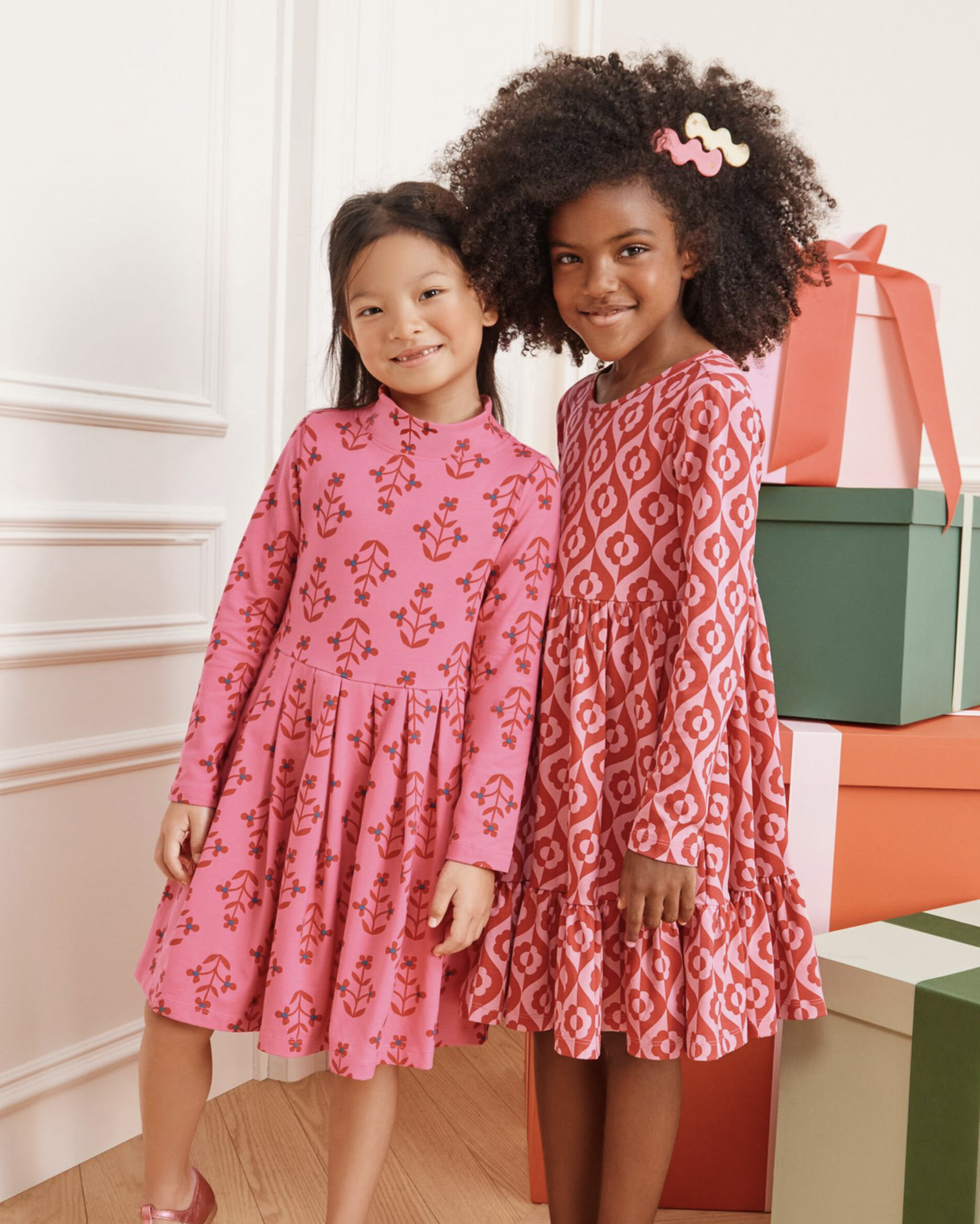 Two young girls standing together wearing long-sleeve patterned dresses from Hanna Andersson, one in a pink floral print and the other in a red geometric print, smiling in a bright room with wrapped gift boxes in the background.