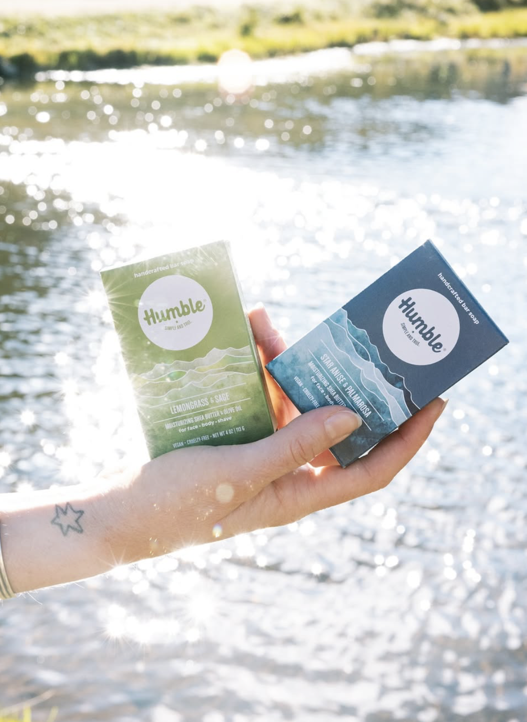 Hand holding two Humble Brands bar soaps beside a sparkling lake, showcasing colorful paper packaging and nature-inspired scents in an outdoor setting.