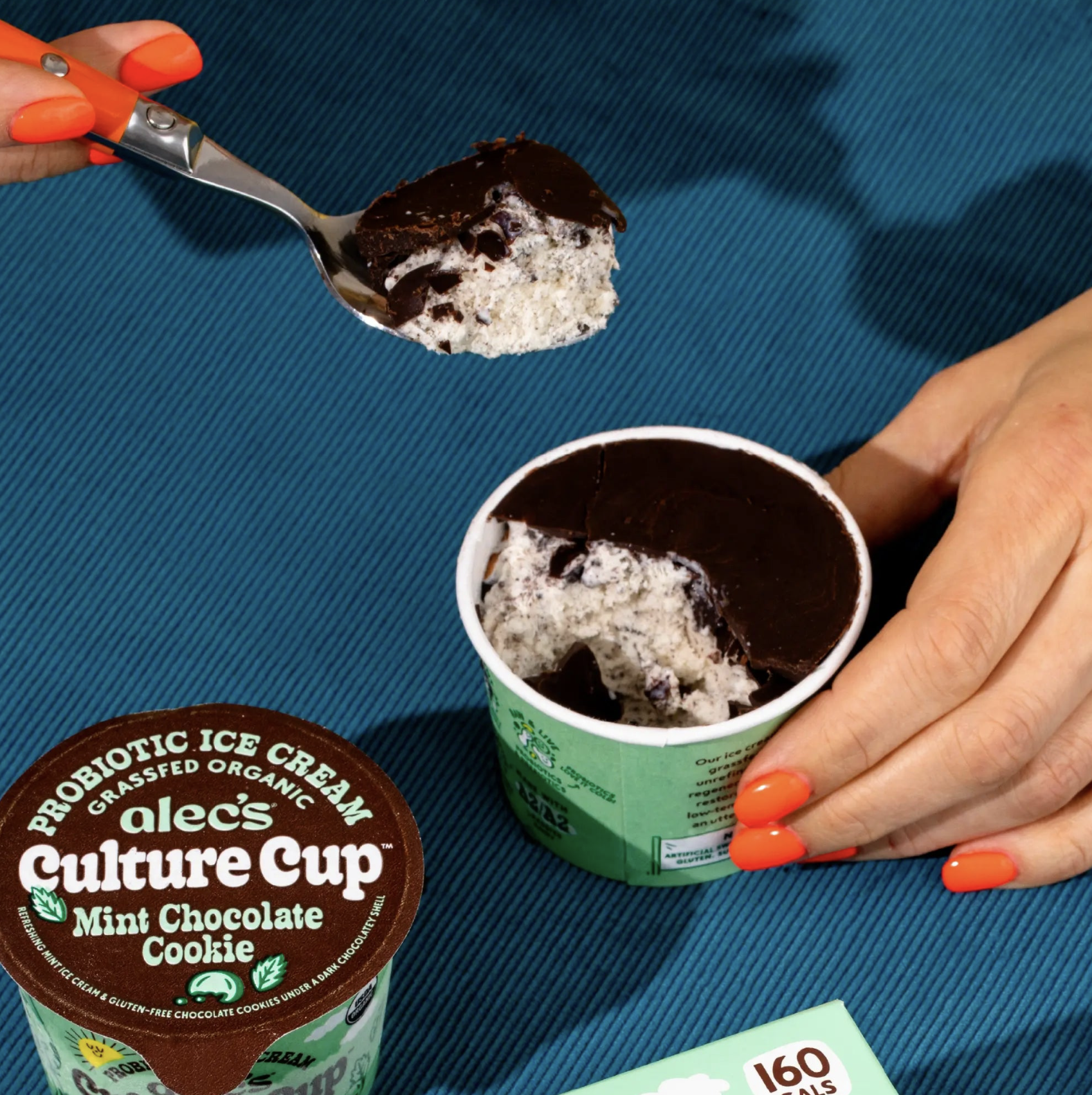 Spoon lifting a bite from an Alec’s Culture Cup Mint Chocolate Cookie probiotic ice cream with a chocolate shell top, held by a hand with bright orange nails against a blue background