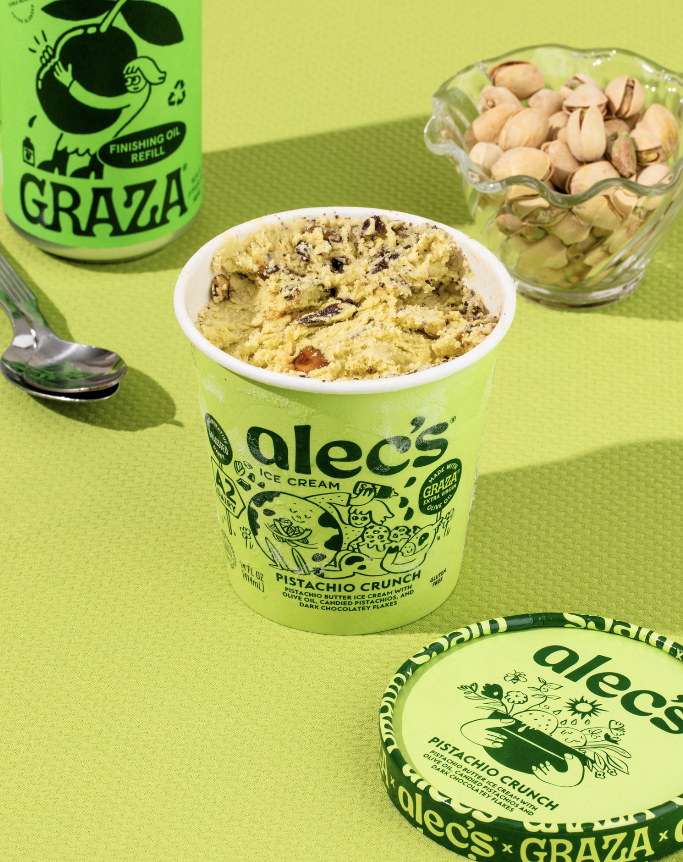 Alec’s Ice Cream Pistachio Crunch pint open on a bright green surface with visible chocolate flakes and nuts, alongside a spoon, a bowl of pistachios and a Graza olive oil bottle in the background