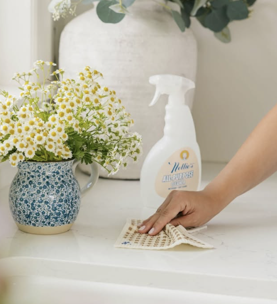 Image 5 A reusable Swedish dishcloth being used to wipe a countertop beside Nellie’s all-purpose cleaner and a vase of fresh flowers.