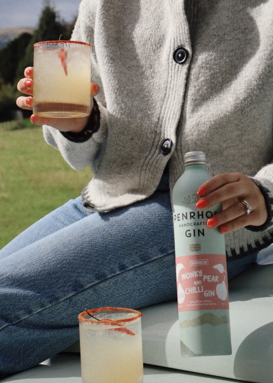 Person holding a cocktail with a chili garnish beside a Penrhos Wonky Pear and Chilli Gin bottle in a recycled aluminium container, photographed outdoors on a sunny day.