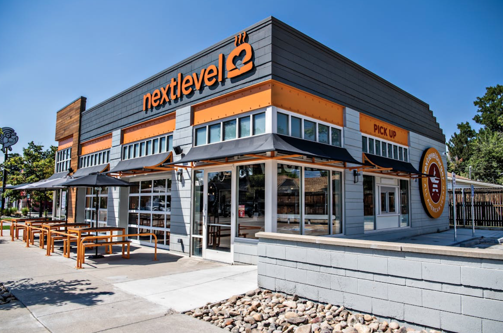 Exterior of a Next Level Veggie Grill restaurant with modern orange and gray signage, outdoor seating and a pick-up window on a sunny day.