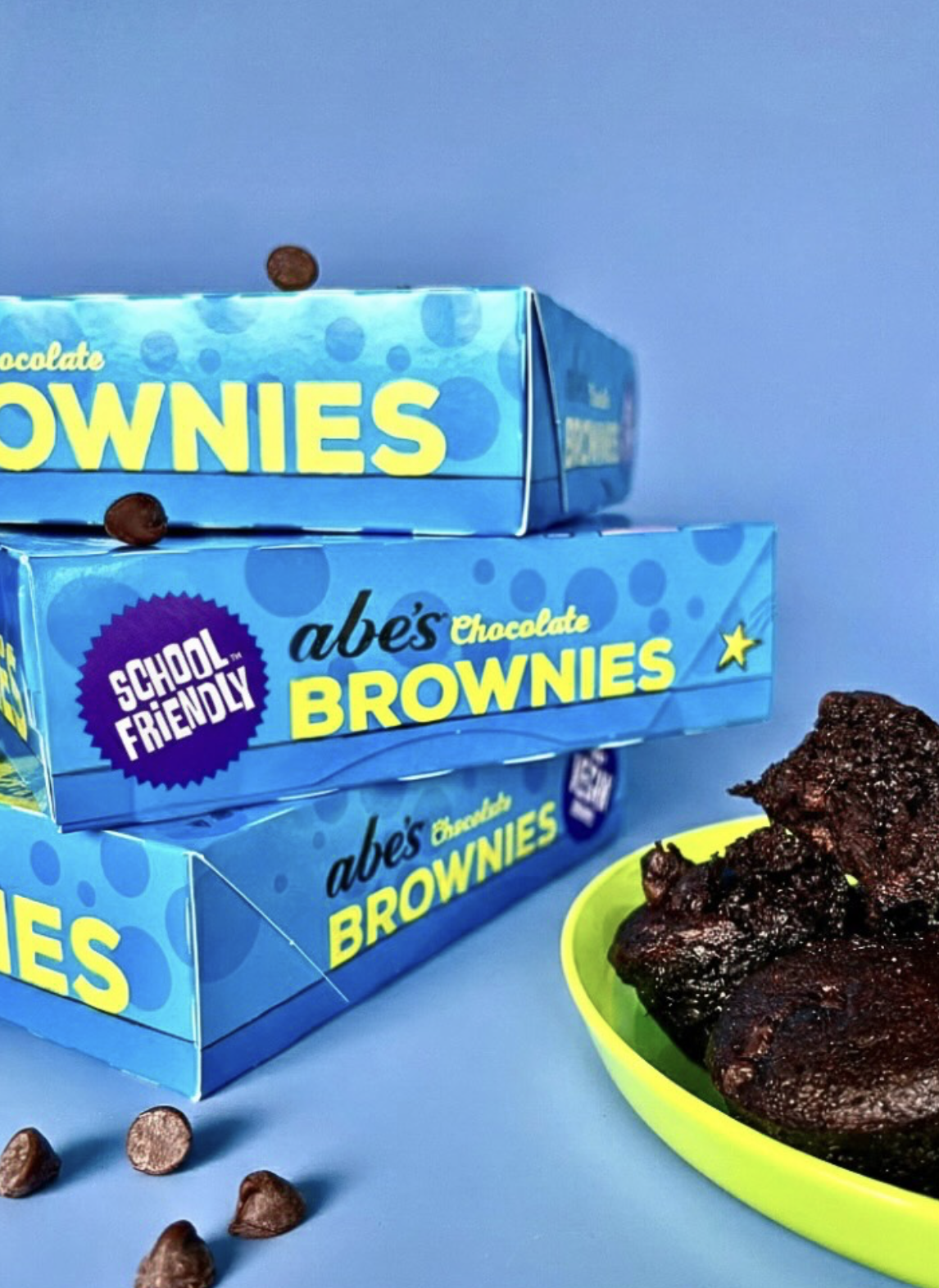 Stacked boxes of Abe’s School-Friendly chocolate brownies beside a plate of rich brownie squares and chocolate chips.