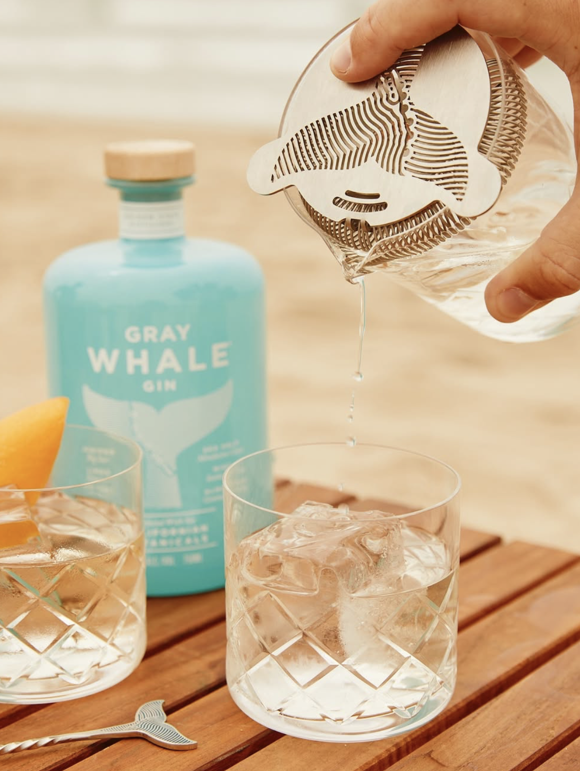 Hand pouring gin through a whale-shaped strainer into a glass with ice, with a bottle of Gray Whale Gin and an orange garnish on a wooden table.