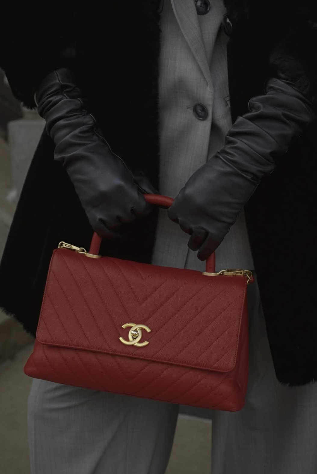 Person wearing a gray suit and black gloves holding a red quilted Chanel handbag with a gold logo.