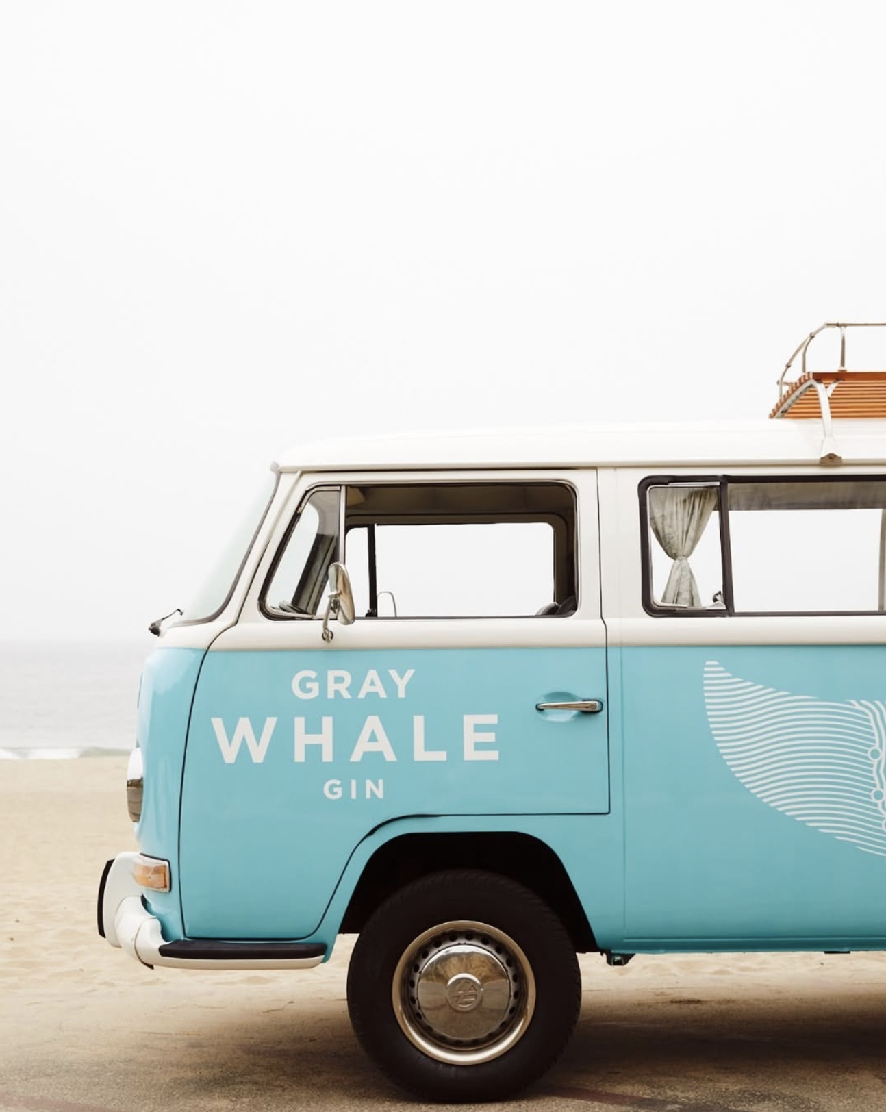 Vintage blue van parked near the beach with Gray Whale Gin branding and whale tail graphic on the side.