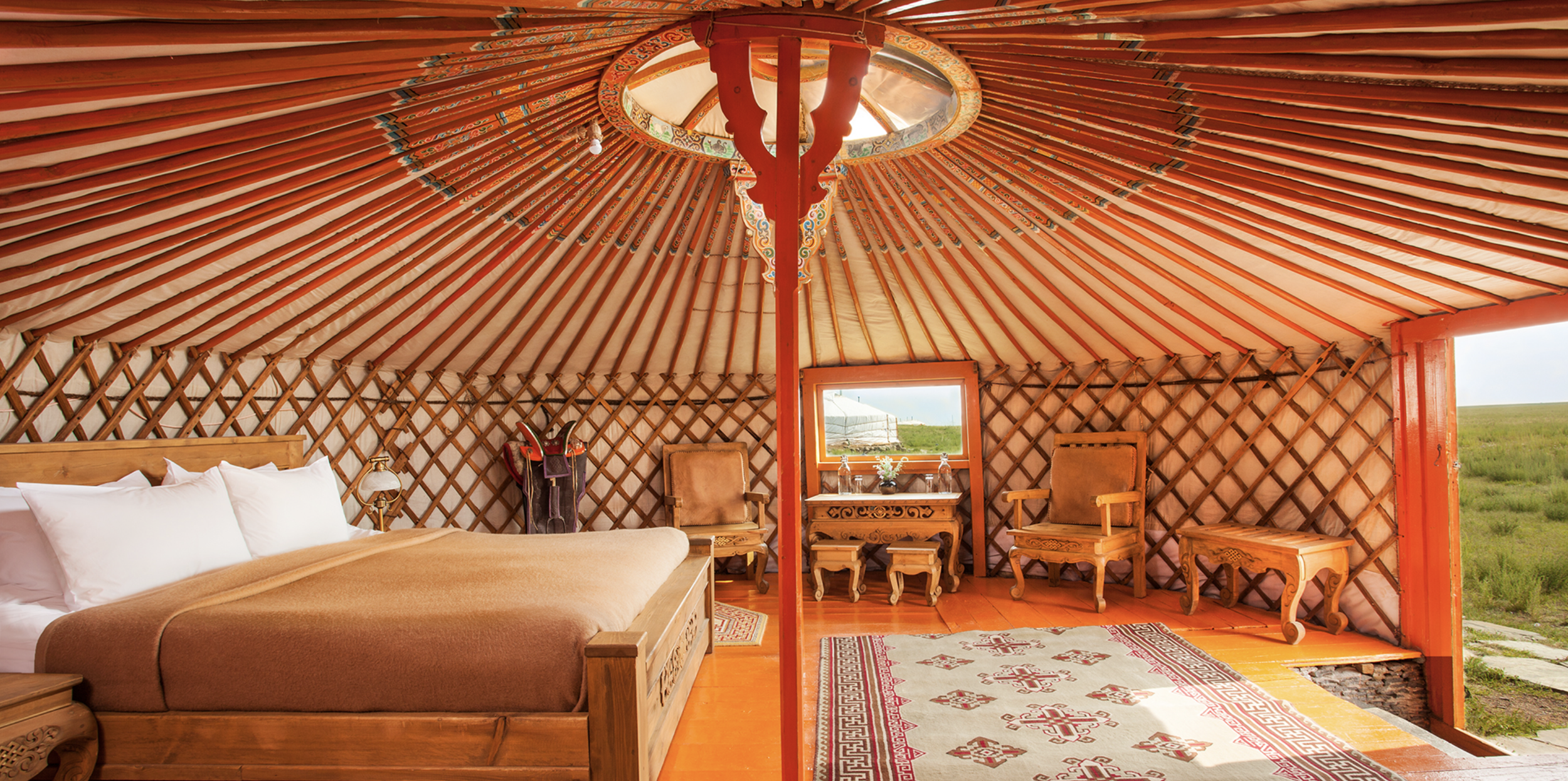 Interior of a traditional Mongolian ger at Three Camel Lodge featuring a wood-framed circular ceiling, carved wooden furniture and a large bed overlooking open grasslands.