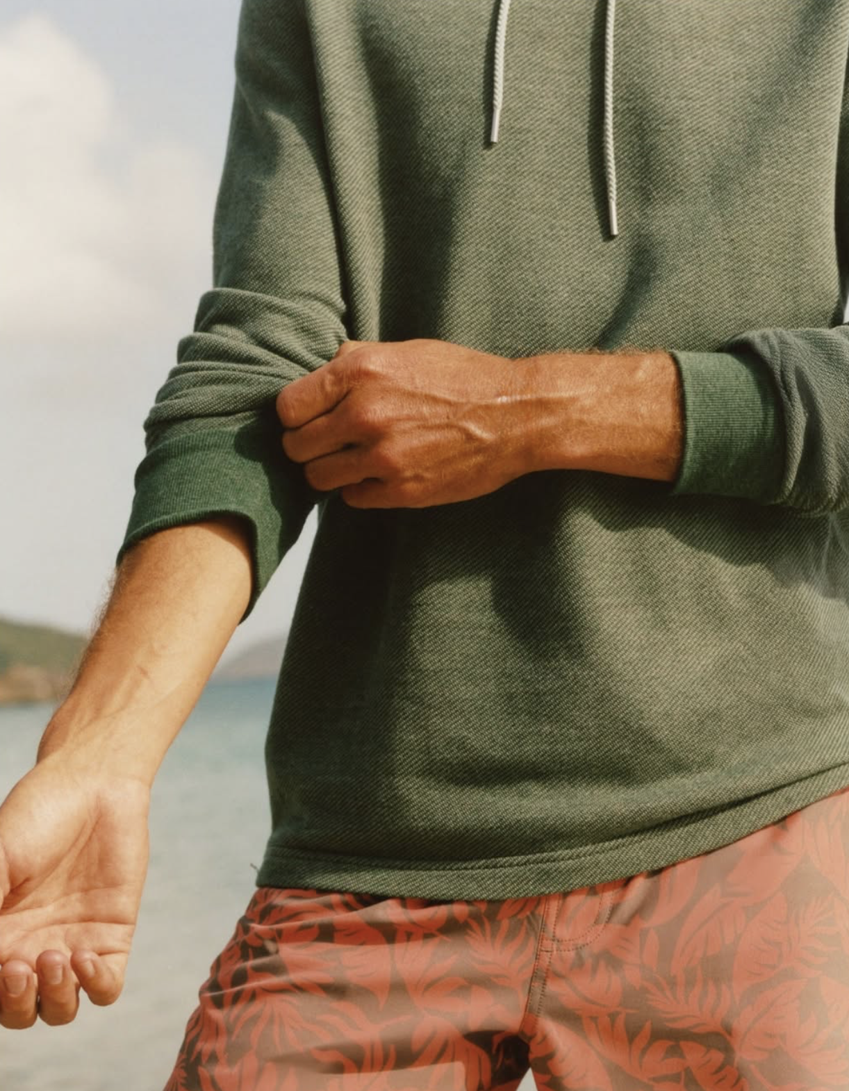 Man wearing a green long-sleeve hoodie with rolled sleeves and patterned swim shorts, standing near the water with a relaxed coastal backdrop.