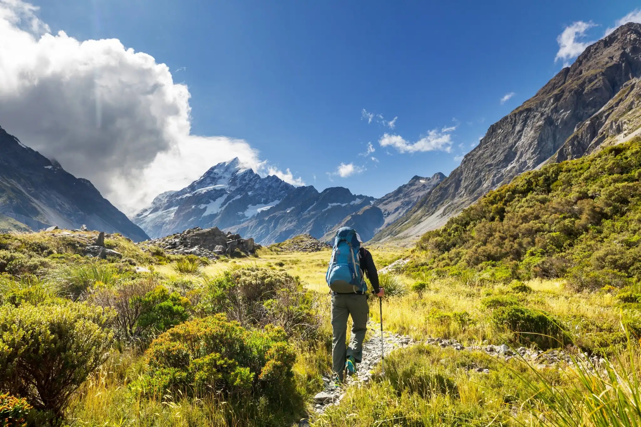 Person hiking on a mountain trail with a backpack, surrounded by grassy alpine landscape and rugged peaks under a bright blue sky