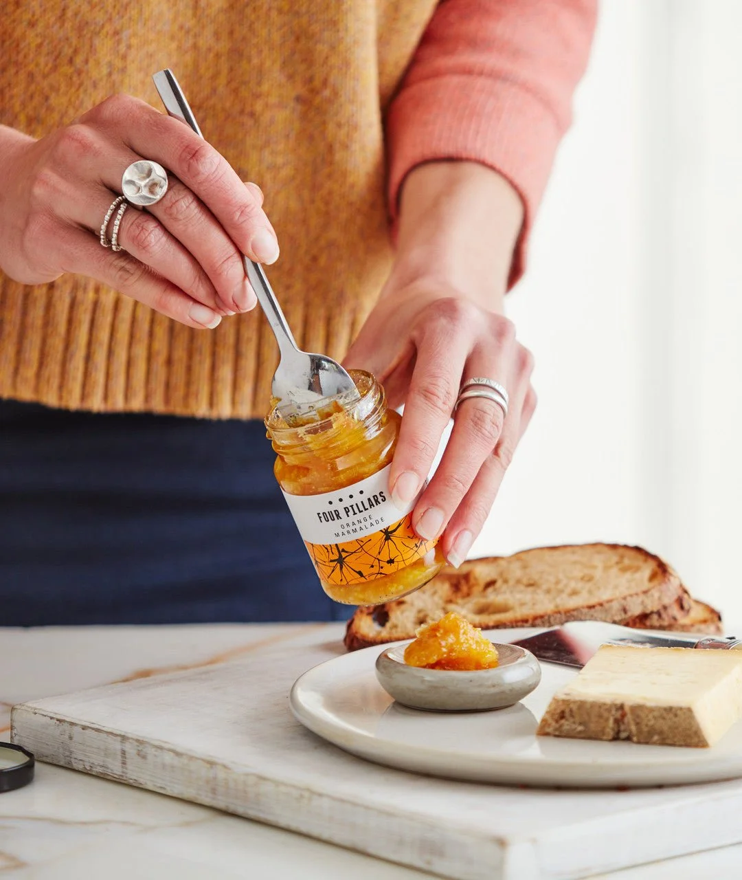 Person scooping Four Pillars Orange Marmalade from a jar onto a plate with bread and cheese on a wooden board