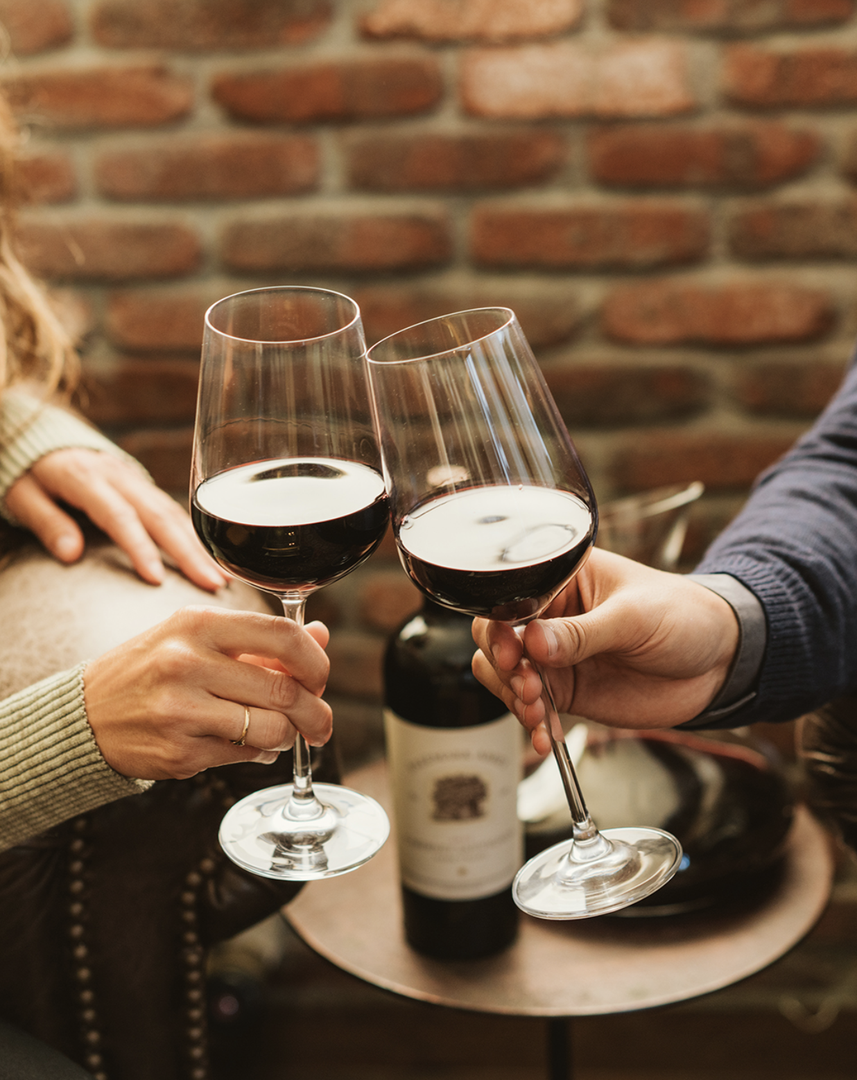 Two people clinking glasses of red wine over a small table with a wine bottle in the background against an exposed brick wall.