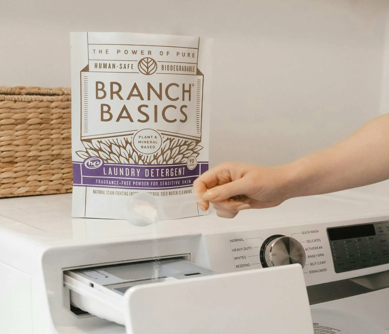 Hand adding Branch Basics laundry detergent powder to a washing machine drawer, highlighting a fragrance-free, human-safe laundry routine