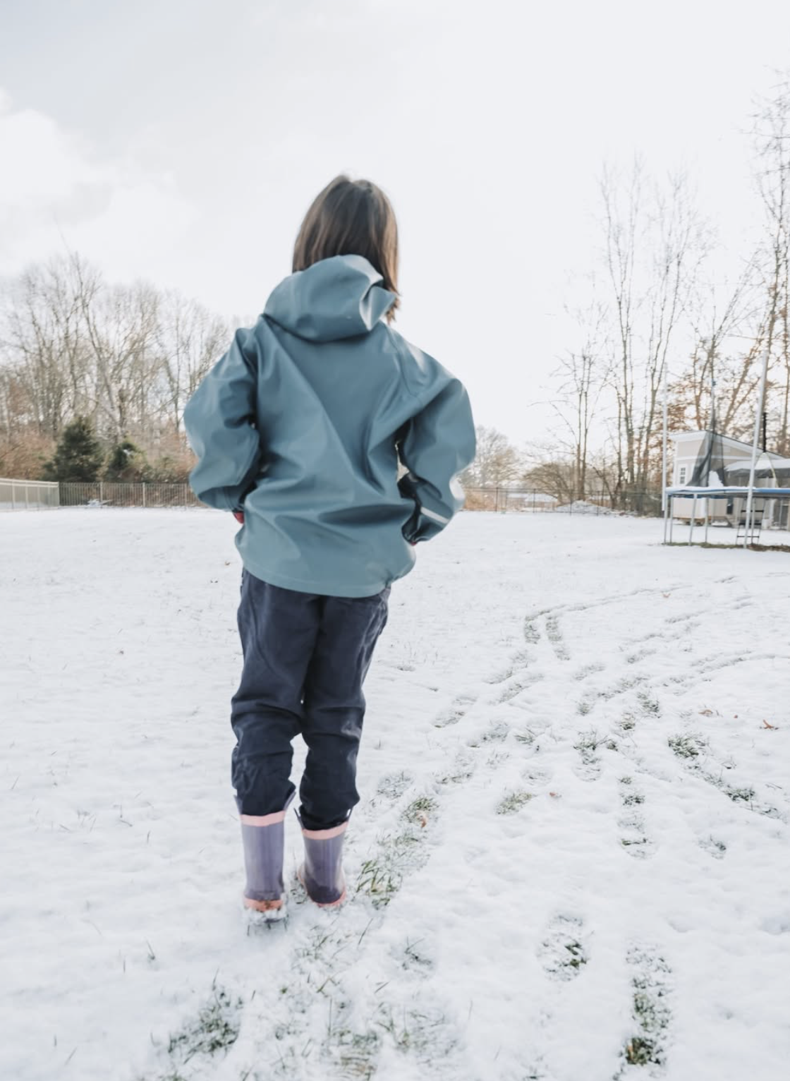 Child walking through a snowy yard wearing a hooded jacket, durable pants and rain boots, highlighting sturdy kids clothing designed for outdoor play in all seasons.