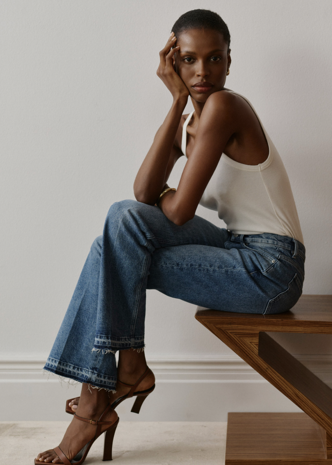 Woman sitting on a wooden table wearing blue DL1961 jeans, styled with a white tank top and heeled sandals in a minimalist studio setting