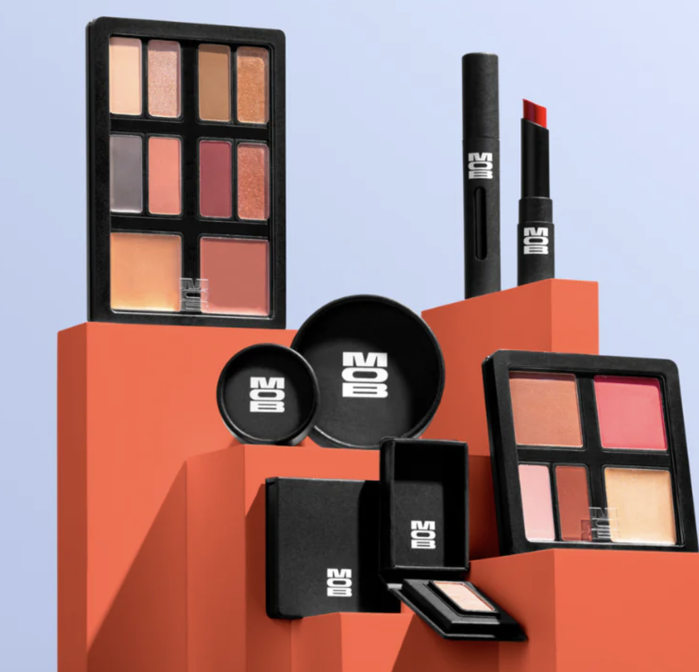 Assortment of MOB Beauty makeup products including refillable palettes, lipsticks and compacts displayed on geometric blocks.