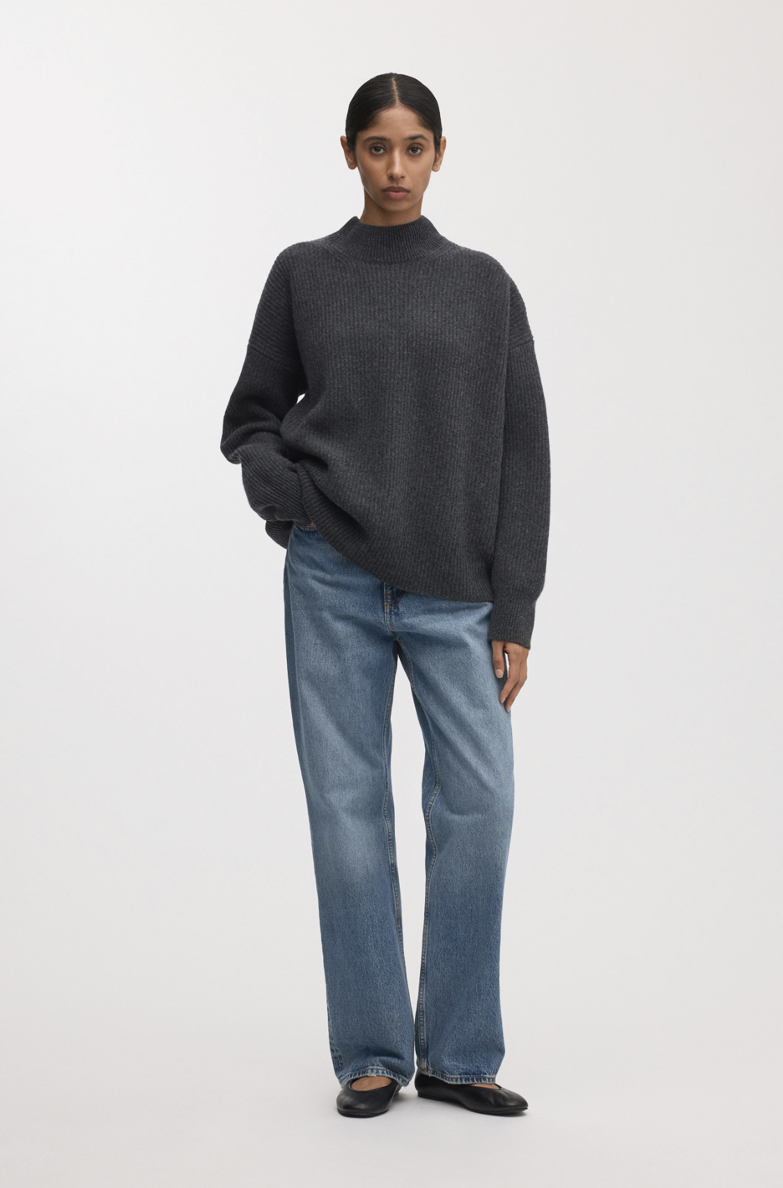 Model wearing a charcoal knit sweater paired with relaxed blue jeans and black flats against a light backdrop.