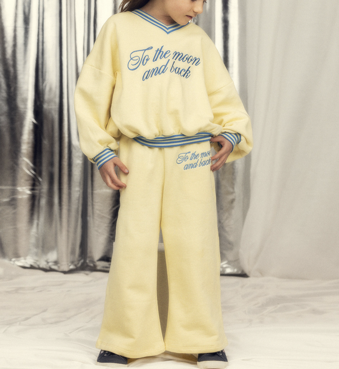 Child wearing a pale yellow matching sweatshirt and wide-leg sweatpants set with blue trim and “To the moon and back” lettering on both pieces against a soft studio backdrop