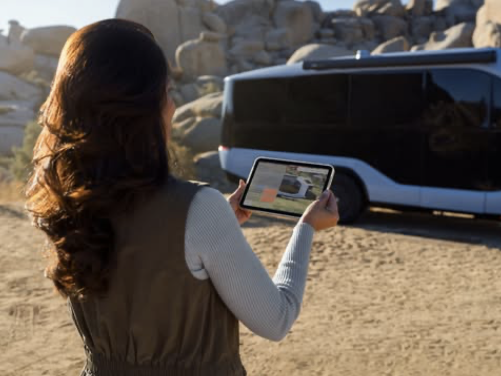 A woman uses a tablet to remotely control the Pebble Flow electric RV trailer, maneuvering it into position at a desert campsite.