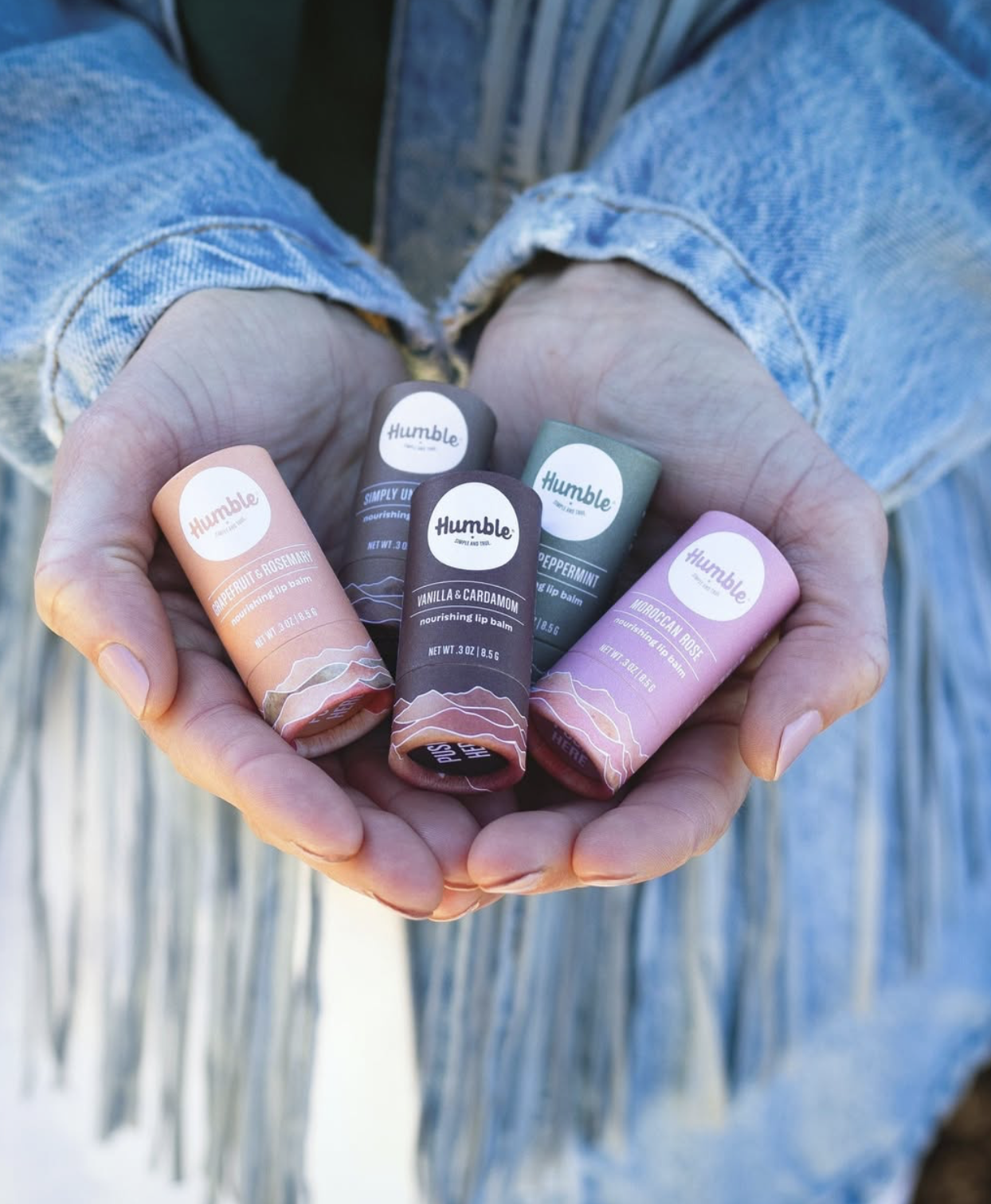 A pair of hands holding several Humble Brands nourishing lip balms in assorted scents, highlighting the brand’s colorful packaging and everyday personal care products.