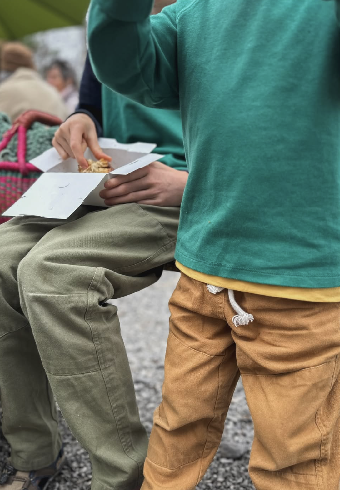 Two children outdoors wearing casual tops and reinforced pants while sharing a snack, showcasing sturdy, everyday kids clothing designed for active use.
