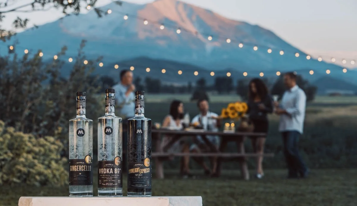 Three Marble Distilling bottles displayed outdoors on a wooden surface with string lights and a mountain landscape in the background while a group gathers around a table behind them.