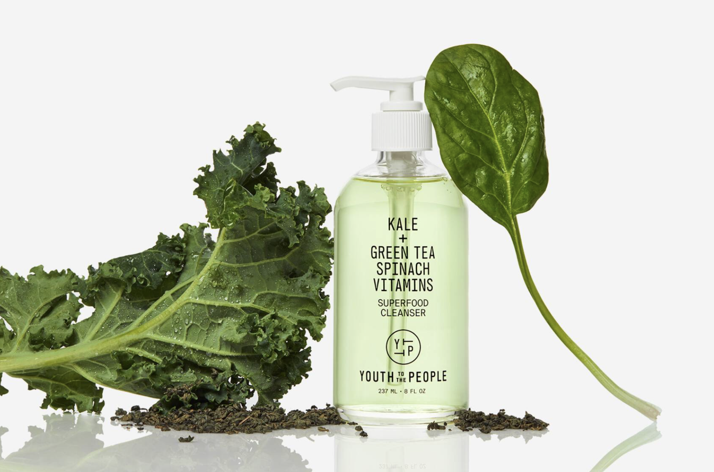 Glass bottle of Youth to the People Kale + Green Tea Spinach Vitamins Superfood Cleanser surrounded by fresh kale leaves, a spinach leaf and loose green tea on a white background.