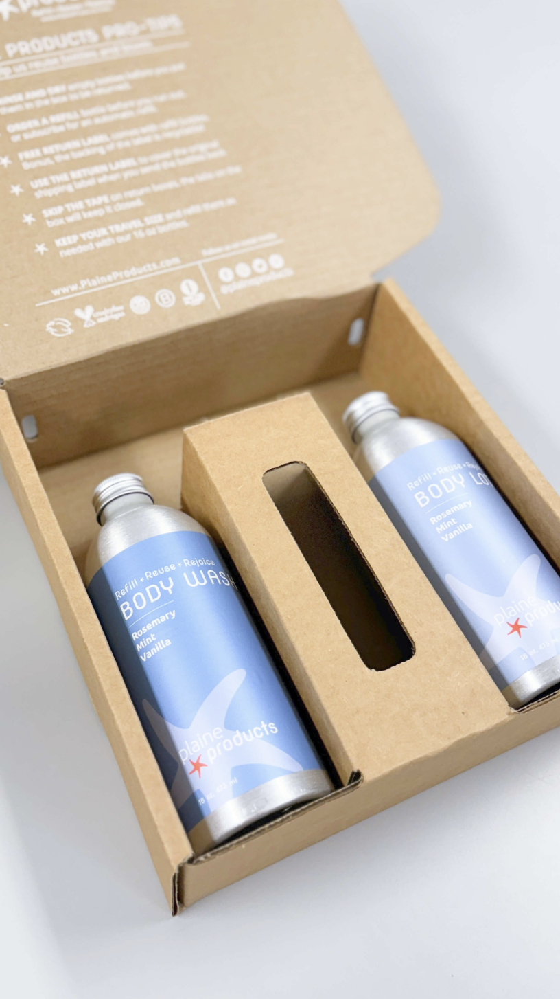 Two refillable aluminum bottles of Plaine Products body wash and body lotion packaged in a cardboard box with insert, emphasizing low-waste design.