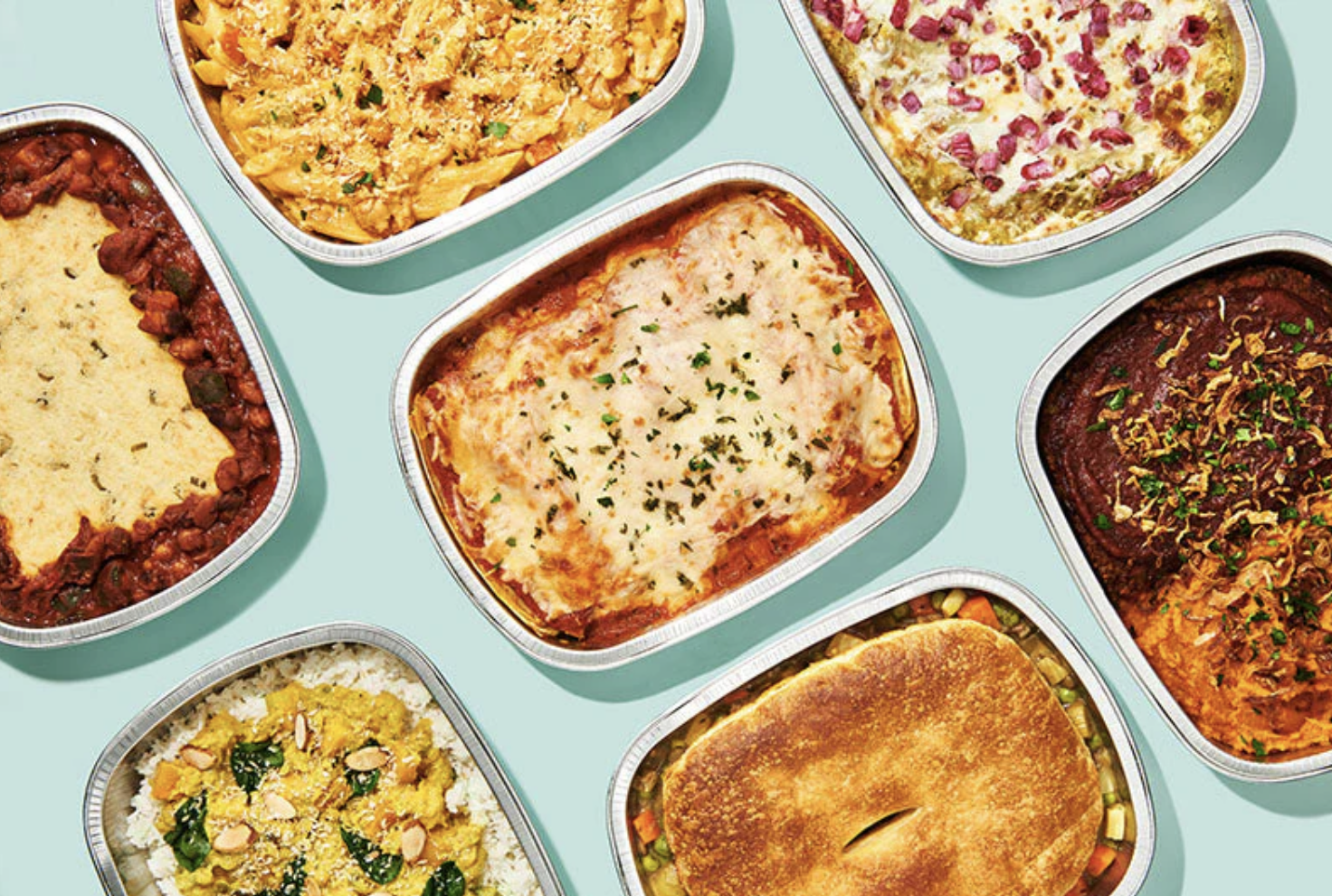 Overhead view of assorted ready-to-eat plant-based meals in rectangular trays, including lasagna, chili, pasta and rice dishes arranged on a light blue background.