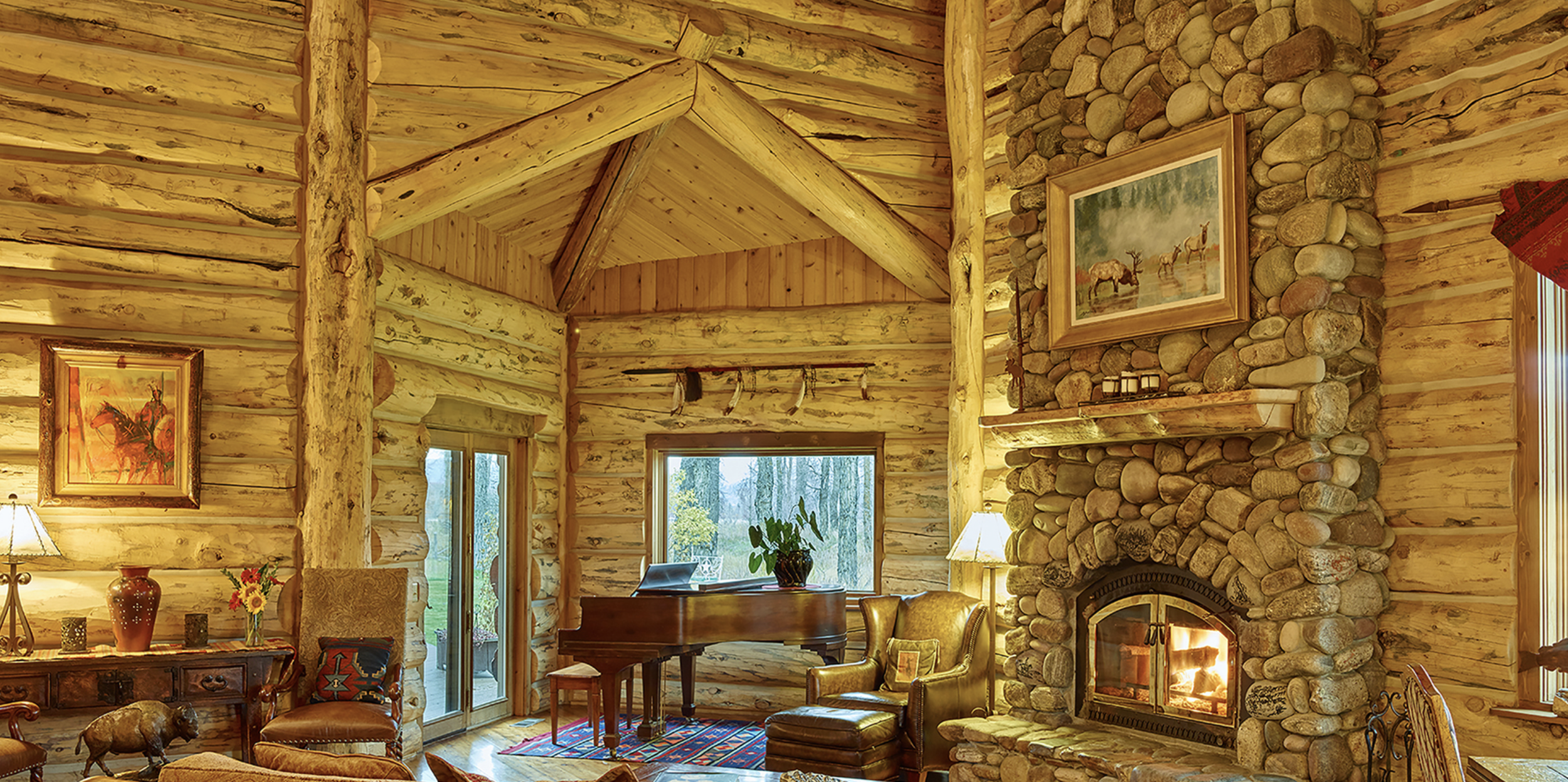 Warm, rustic interior of a log cabin at The Ranch at Rock Creek in Montana with a grand piano and a large stone fireplace glowing with firelight.