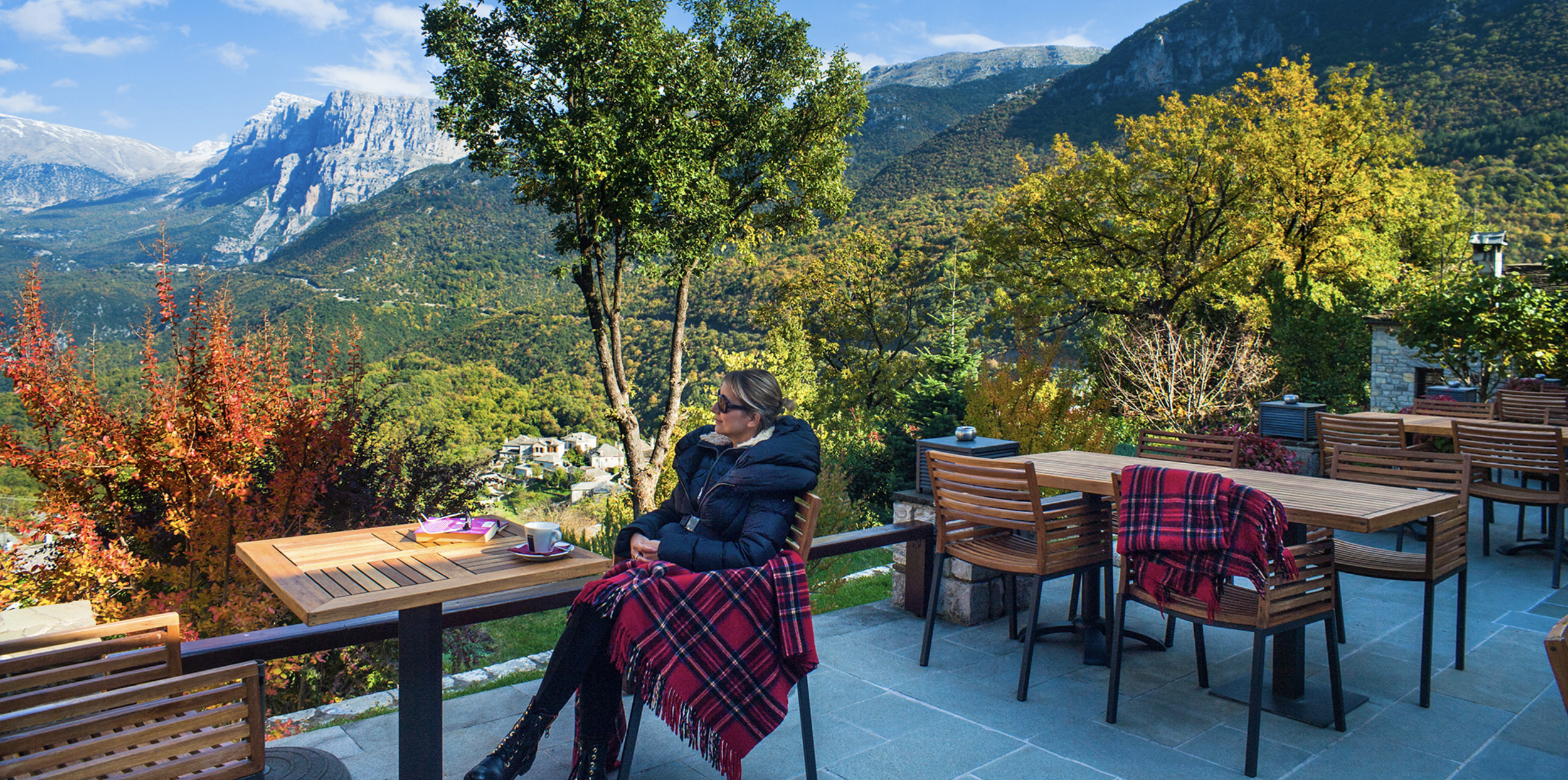 Guest seated at an outdoor terrace at Aristi Mountain Resort in Greece, overlooking dramatic mountain scenery in autumn with trees in vibrant gold and red.