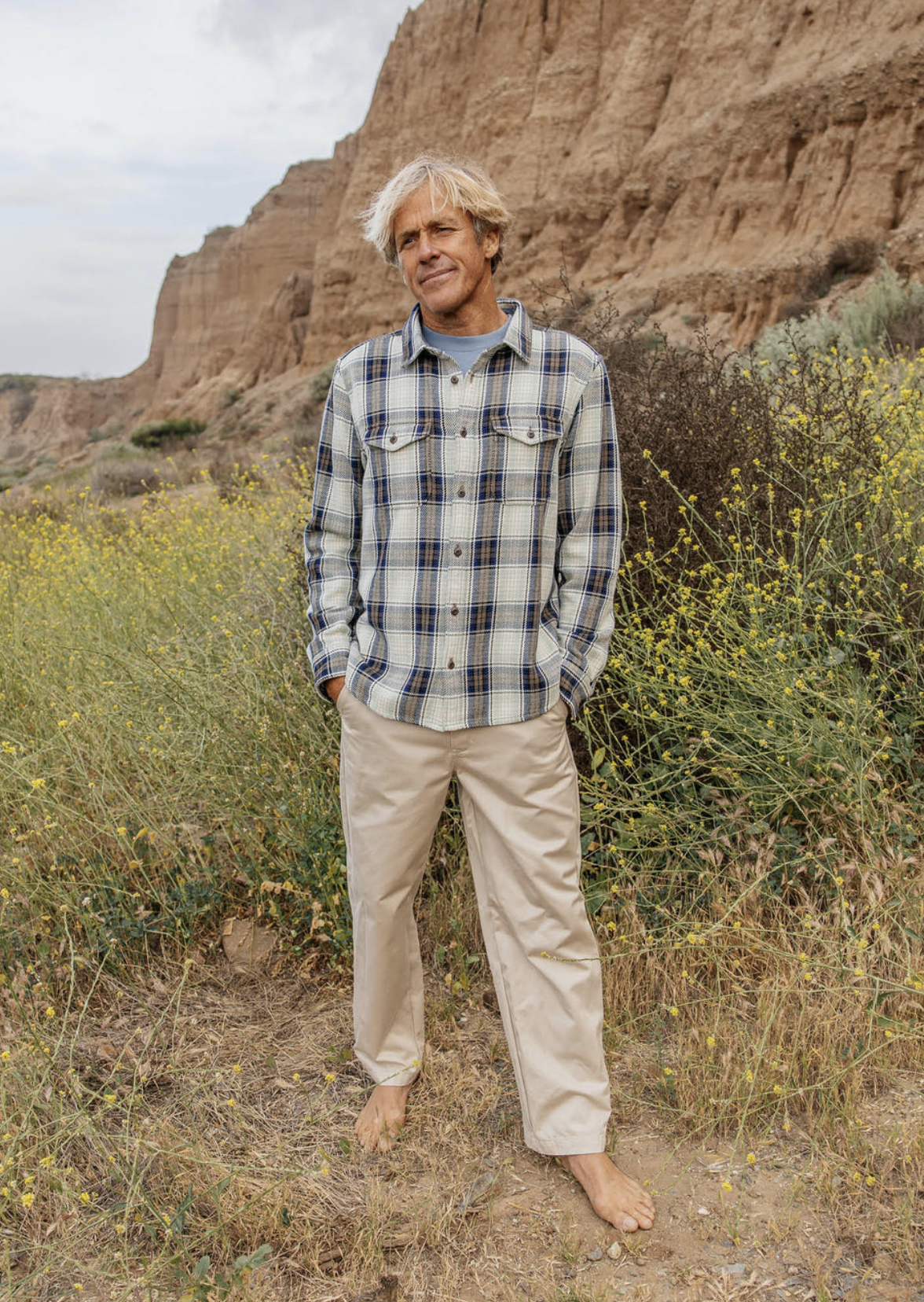 Person wearing a light plaid flannel shirt and tan pants standing barefoot on a dirt path with rocky cliffs and yellow wildflowers in the background.