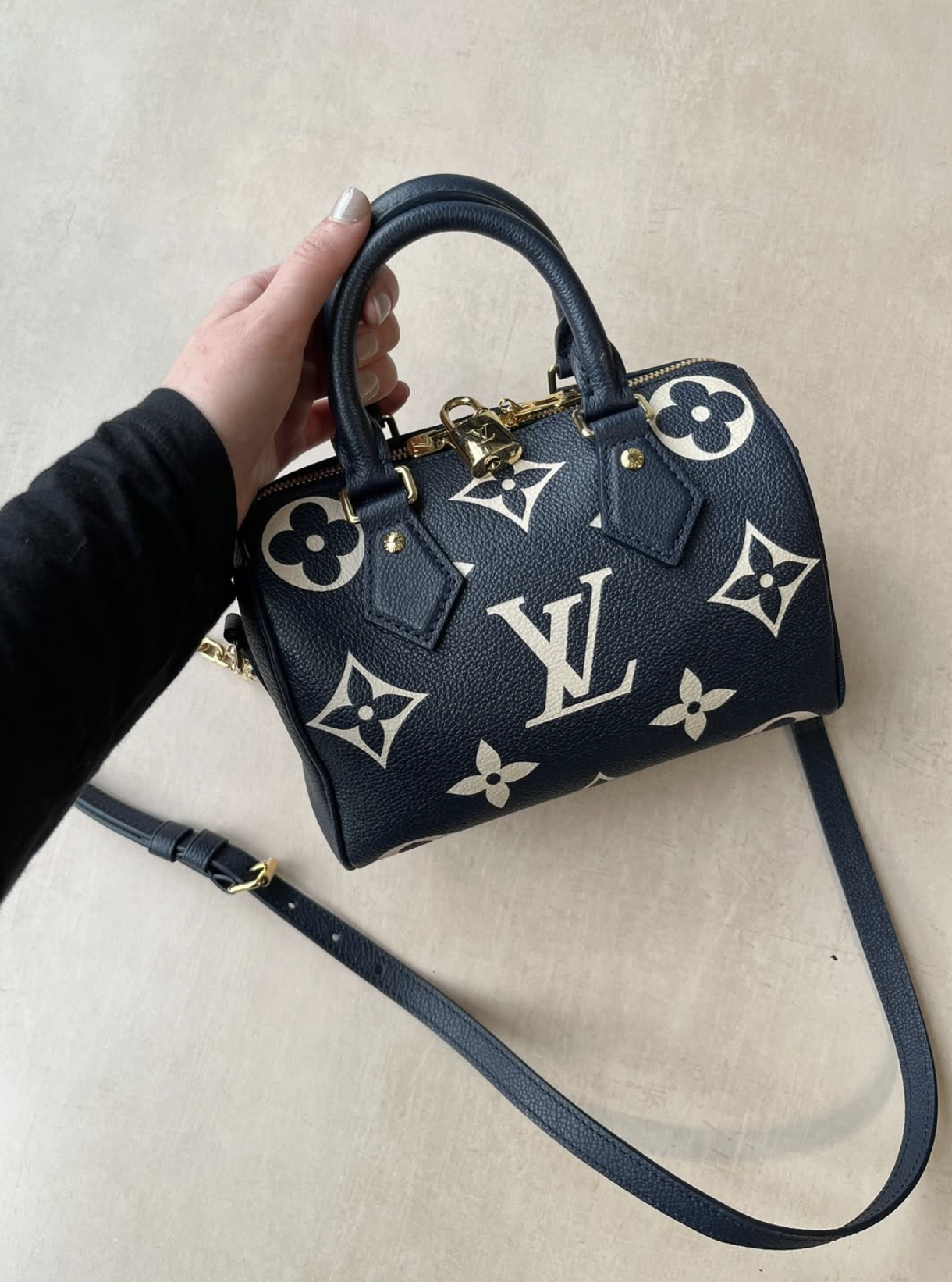 Hand holding a navy Louis Vuitton handbag with oversized monogram pattern and a detachable shoulder strap.