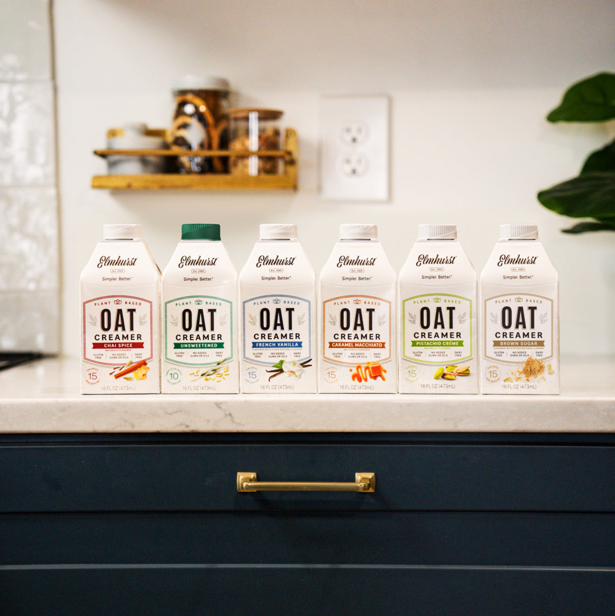 Six Elmhurst Oat Creamer cartons lined up on a kitchen counter, featuring flavors like Chai Spice, Unsweetened, French Vanilla, Caramel Macchiato, Pistachio Crème and Brown Sugar Vanilla.