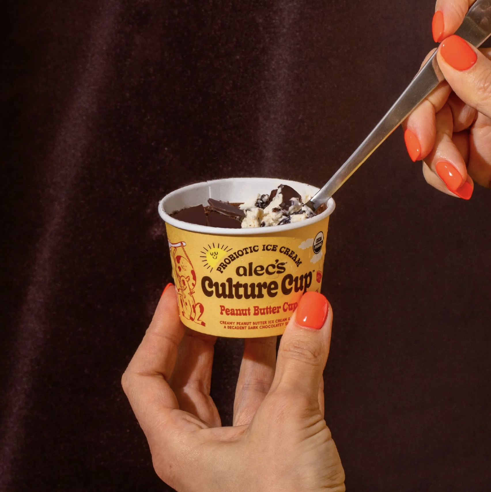 Hand holding an Alec’s Culture Cup probiotic ice cream in Peanut Butter Cup flavor while scooping into the chocolate shell with a spoon against a dark background