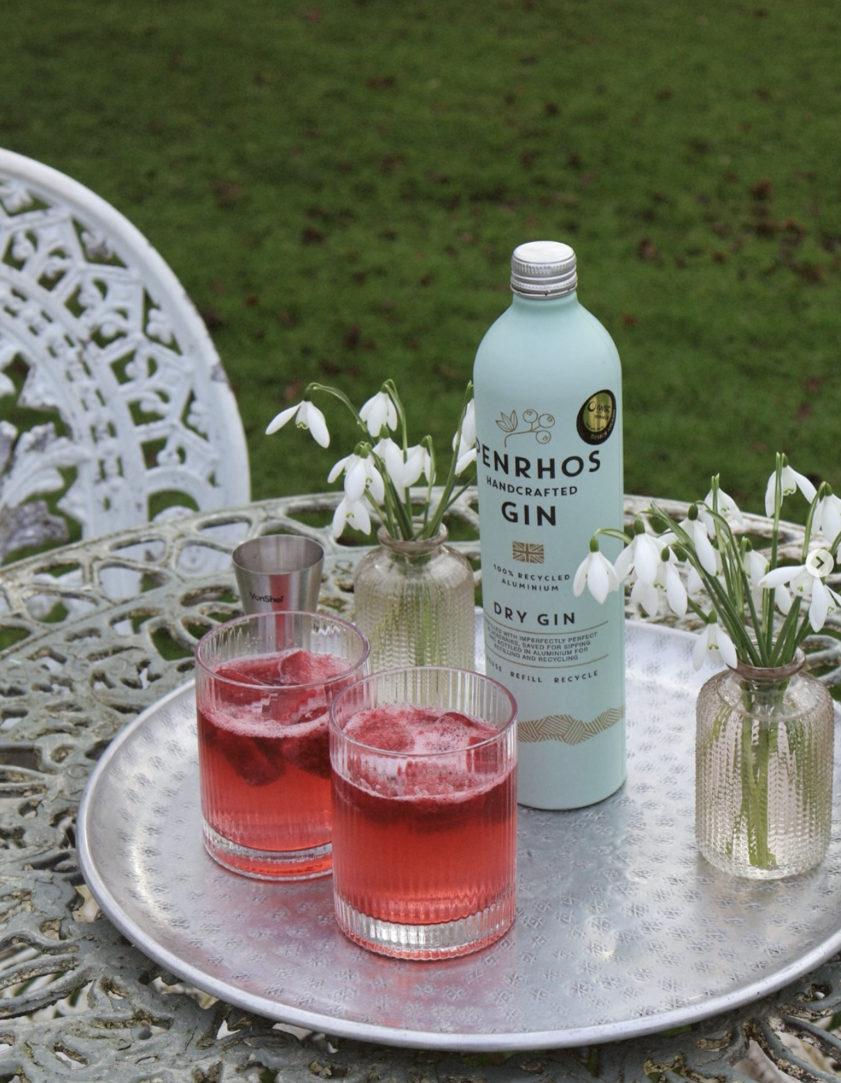 Penrhos Dry Gin in a recycled aluminium bottle displayed on an outdoor table with two red gin cocktails and small vases of white flowers on a silver tray.