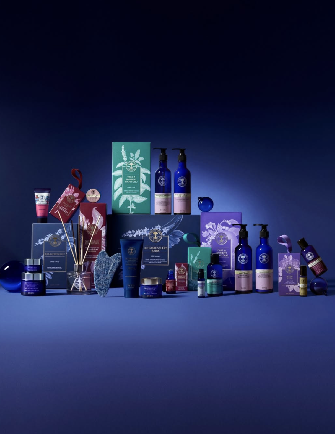 A curated display of Neal’s Yard Remedies skincare, bath and body and aromatherapy products arranged against a deep blue background, featuring cobalt blue bottles, botanical packaging and gift sets.