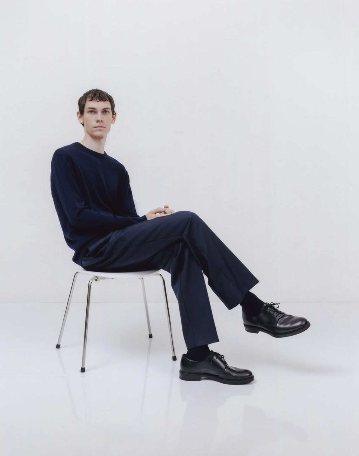 Model wearing a navy long-sleeve knit top with matching tailored navy trousers and black leather lace-up shoes, seated on a minimalist metal chair against a neutral background.