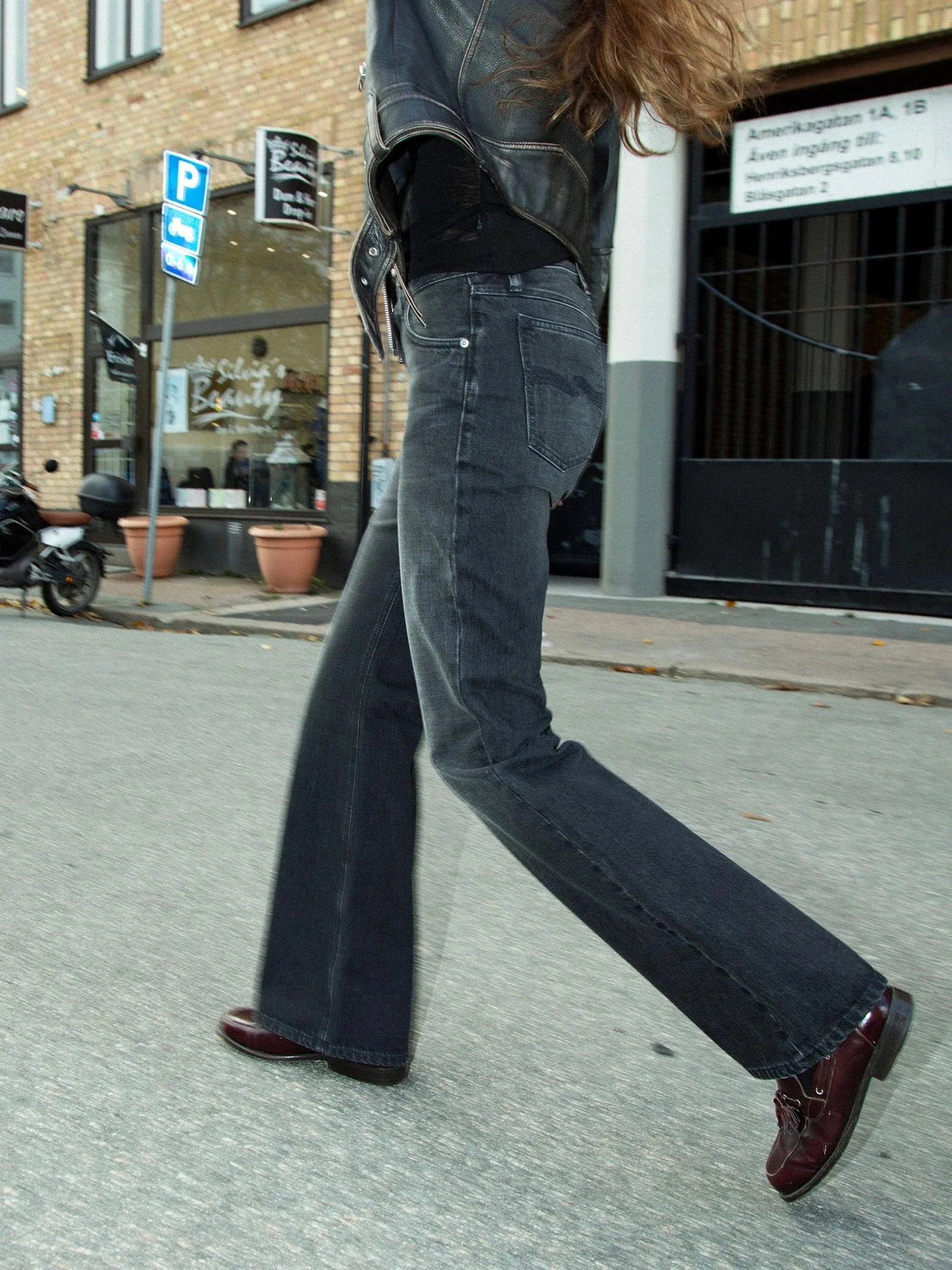 Person walking down a city street wearing faded black Nudie Jeans with a wide-leg fit, styled with a black top, leather jacket and dark brown shoes