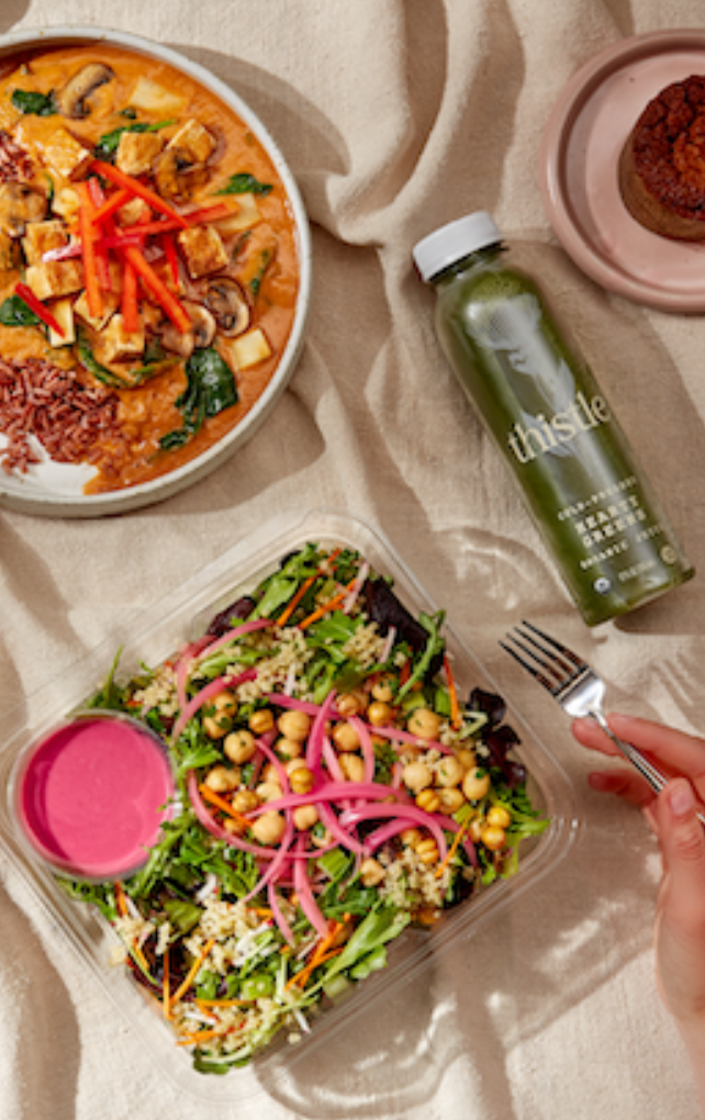 Top view of plant-based prepared meals including a colorful salad with chickpeas and pickled onions, a curry bowl with vegetables and rice, and a green juice bottle.