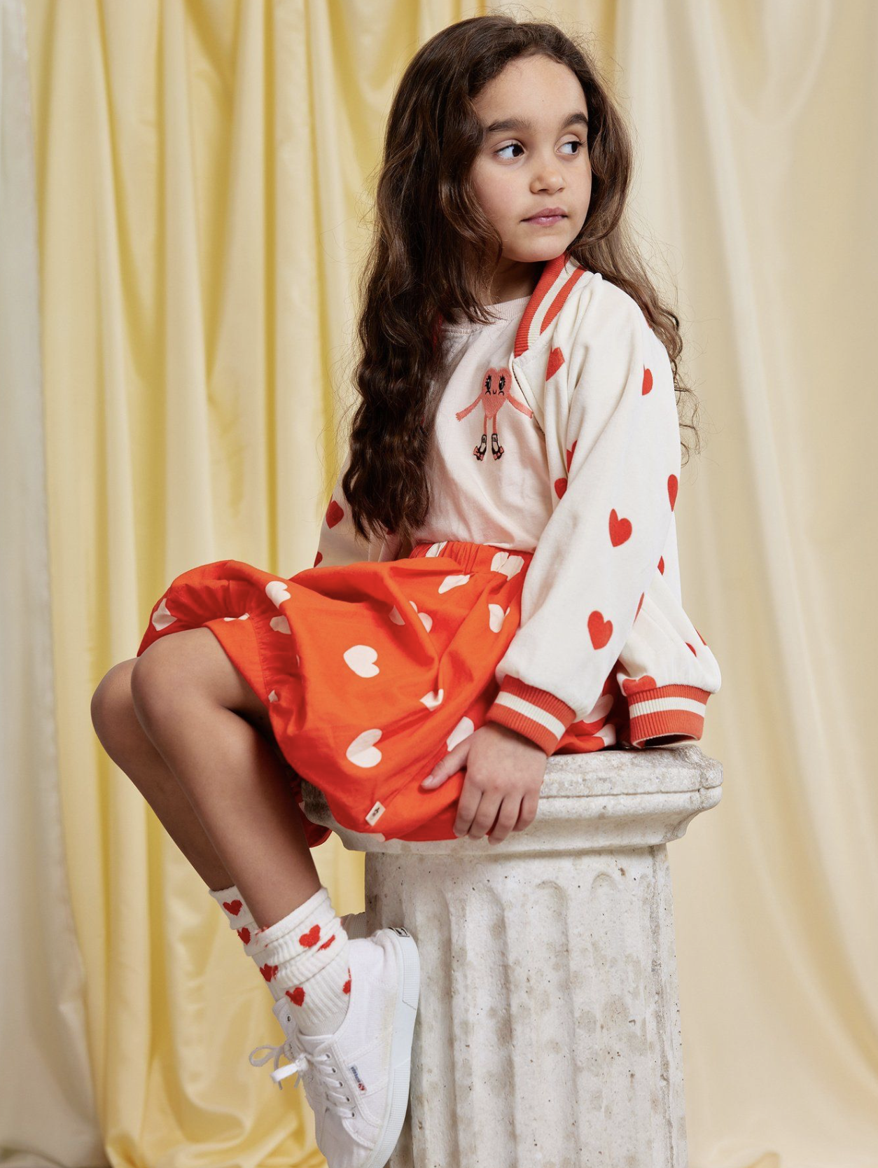 A young girl sitting on a stone pedestal wearing a cream and red heart-patterned jacket, a matching red skirt with heart prints and white sneakers, against a soft yellow draped backdrop.